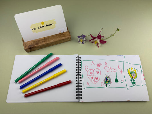 Open notebook with drawings and coloured pens on a light green surface for Being Me with Friends Kit by Wild Hearts