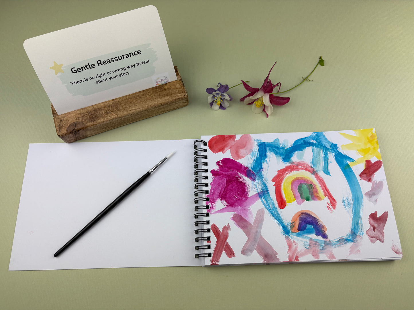 Open sketchbook showing a colourful child’s painting inspired by the My Story My Way and Two Homes One Me Identity creative support kit, with prompt card, paintbrush and wooden card holder displayed on a sage green background.