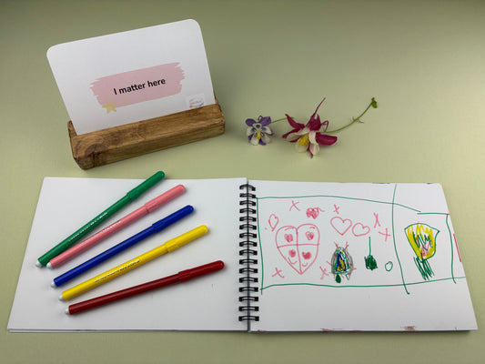 Reassurance card for children struggling with feelings and self esteem at home and school. Creative Kit, with open sketchbook with drawings, coloured Giotto pens, and a card on a green surface. I matter here by Wild Hearts Creative