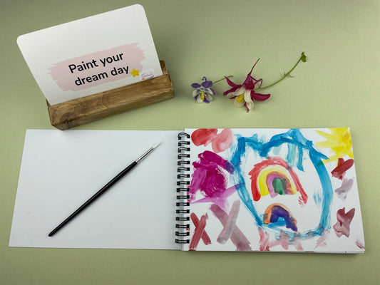 Open sketchbook showing a colourful child’s painting inspired by the Paint Your Confidence creative support kit, with prompt card, paintbrush and wooden card holder displayed on a sage green background.