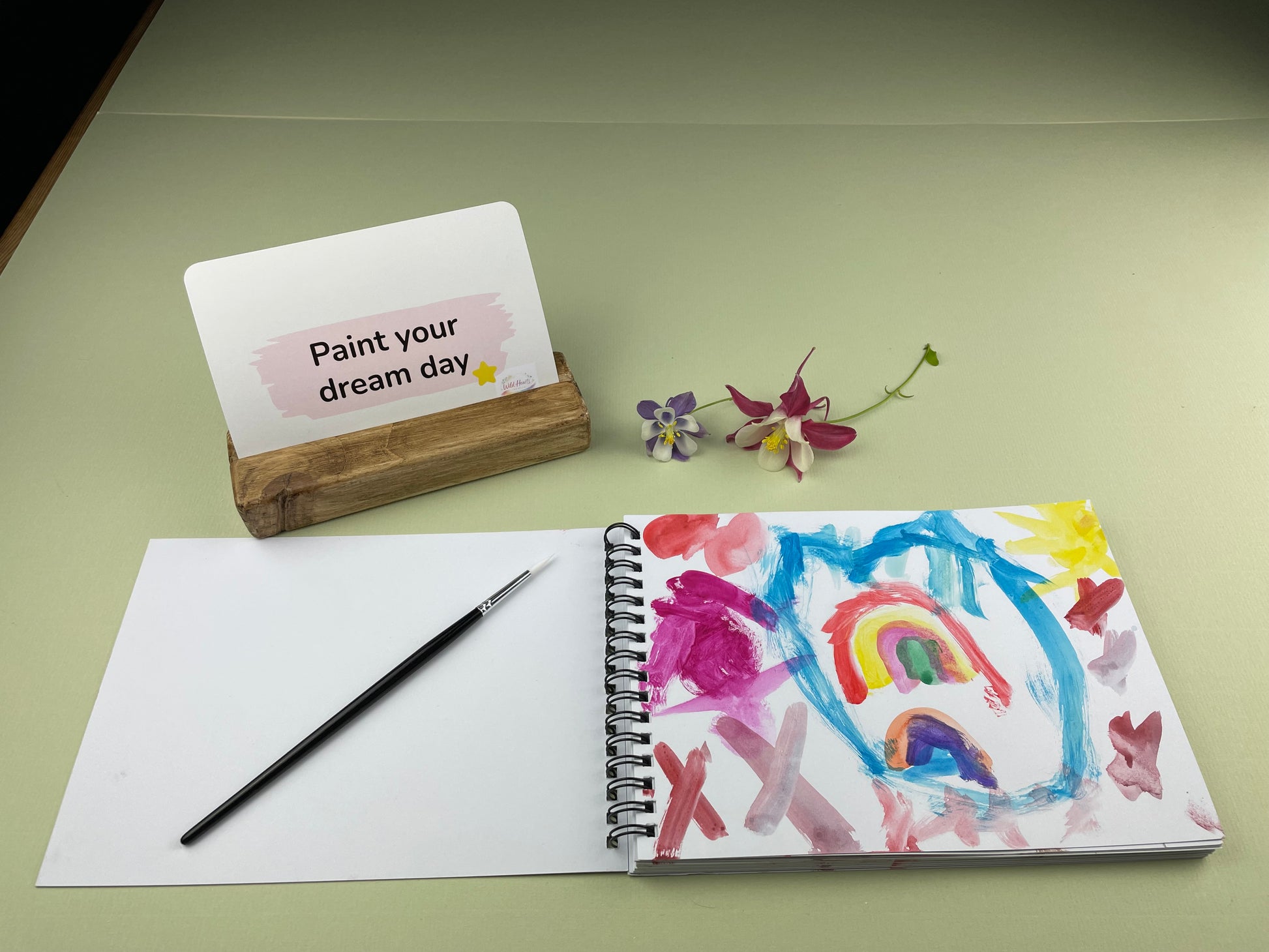 Open sketchbook showing a colourful child’s painting inspired by the Paint Your Confidence creative support kit, with prompt card, paintbrush and wooden card holder displayed on a sage green background.