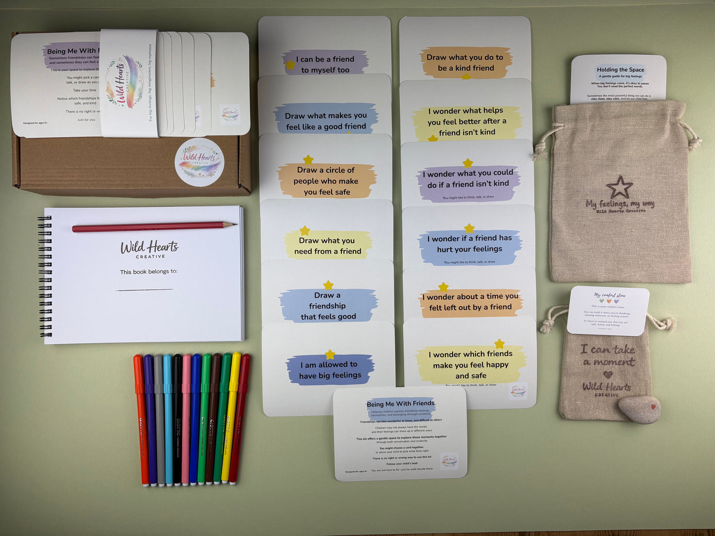 Flat lay of the Being Me With Friends support kit for children, including affirmation cards, creative prompt cards, sketchbook, paints, comfort stone and drawstring pouch.