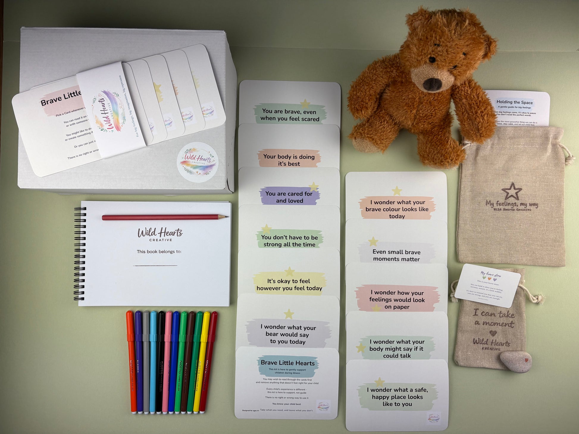 Flat lay of the Hospital and Illness support kit for children, including affirmation cards, creative prompt cards, sketchbook, paints, comfort stone and drawstring pouch.