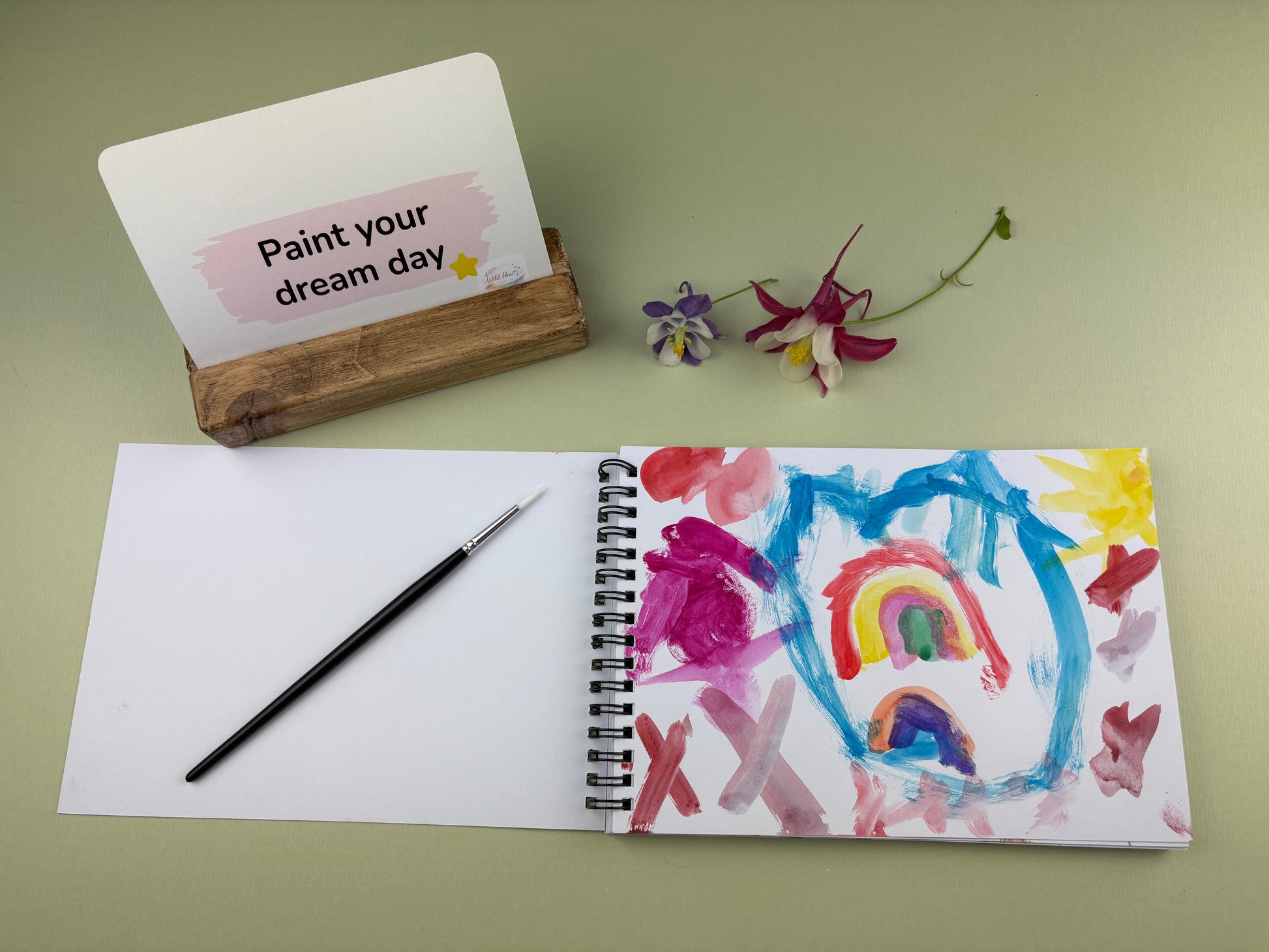 Open sketchbook showing a colourful child’s painting inspired by the Paint Your Confidence creative support kit, with prompt card, paintbrush and wooden card holder displayed on a sage green background.