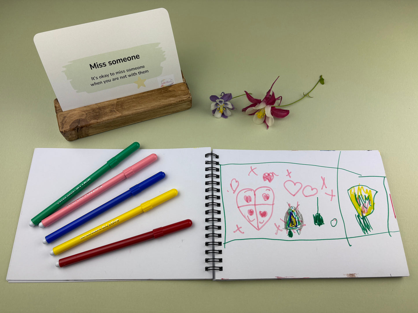 Reassurance it's okay to miss someone card for children when going through change.  Creative Kit, with open sketchbook with drawings, coloured Giotto pens, and a card on a green surface.  Divorce and Separation Kit for missing absent parent by Wild Hearts Creative