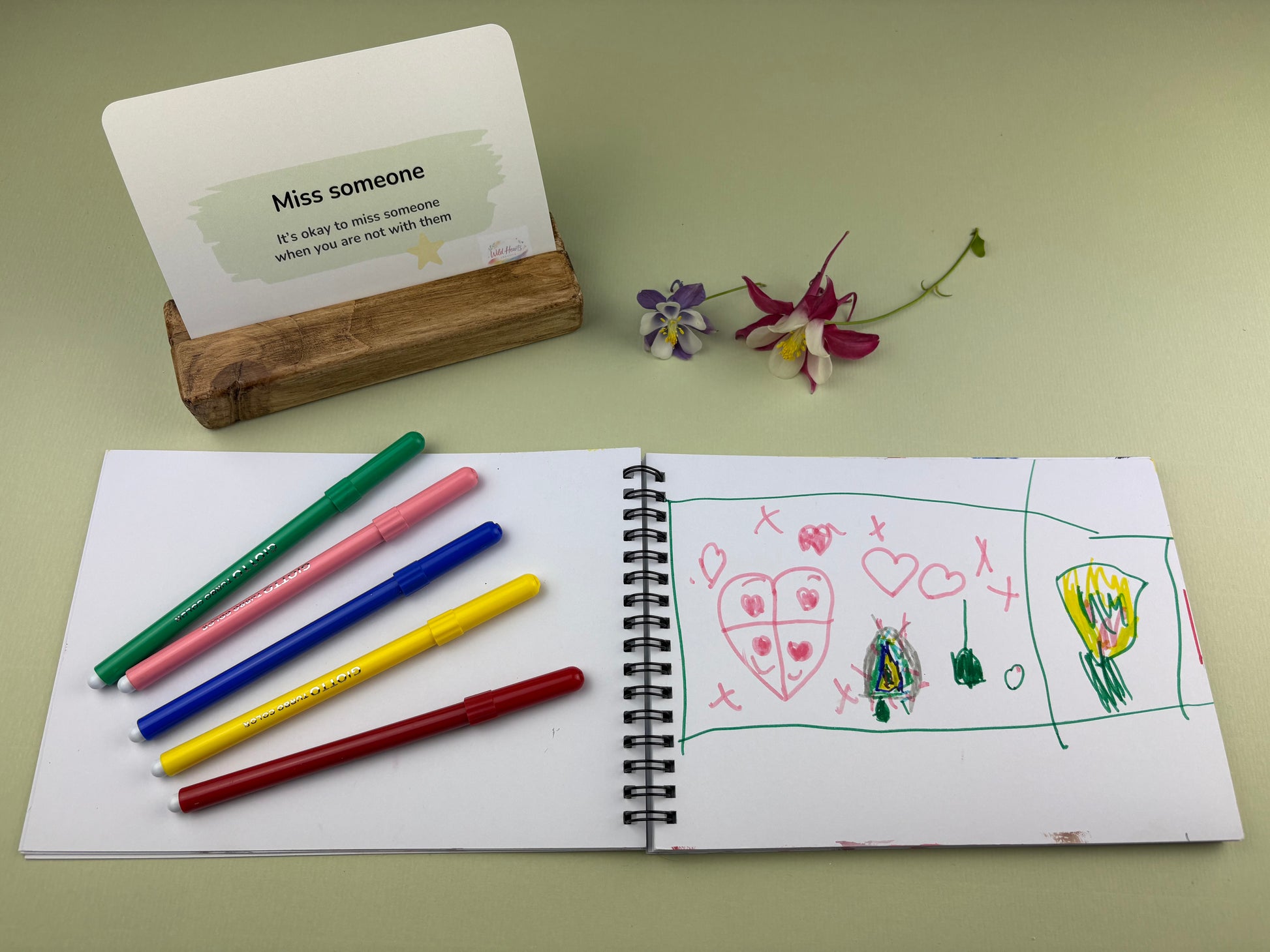 Reassurance it's okay to miss someone card for children when going through change.  Creative Kit, with open sketchbook with drawings, coloured Giotto pens, and a card on a green surface.  Divorce and Separation Kit for missing absent parent by Wild Hearts Creative