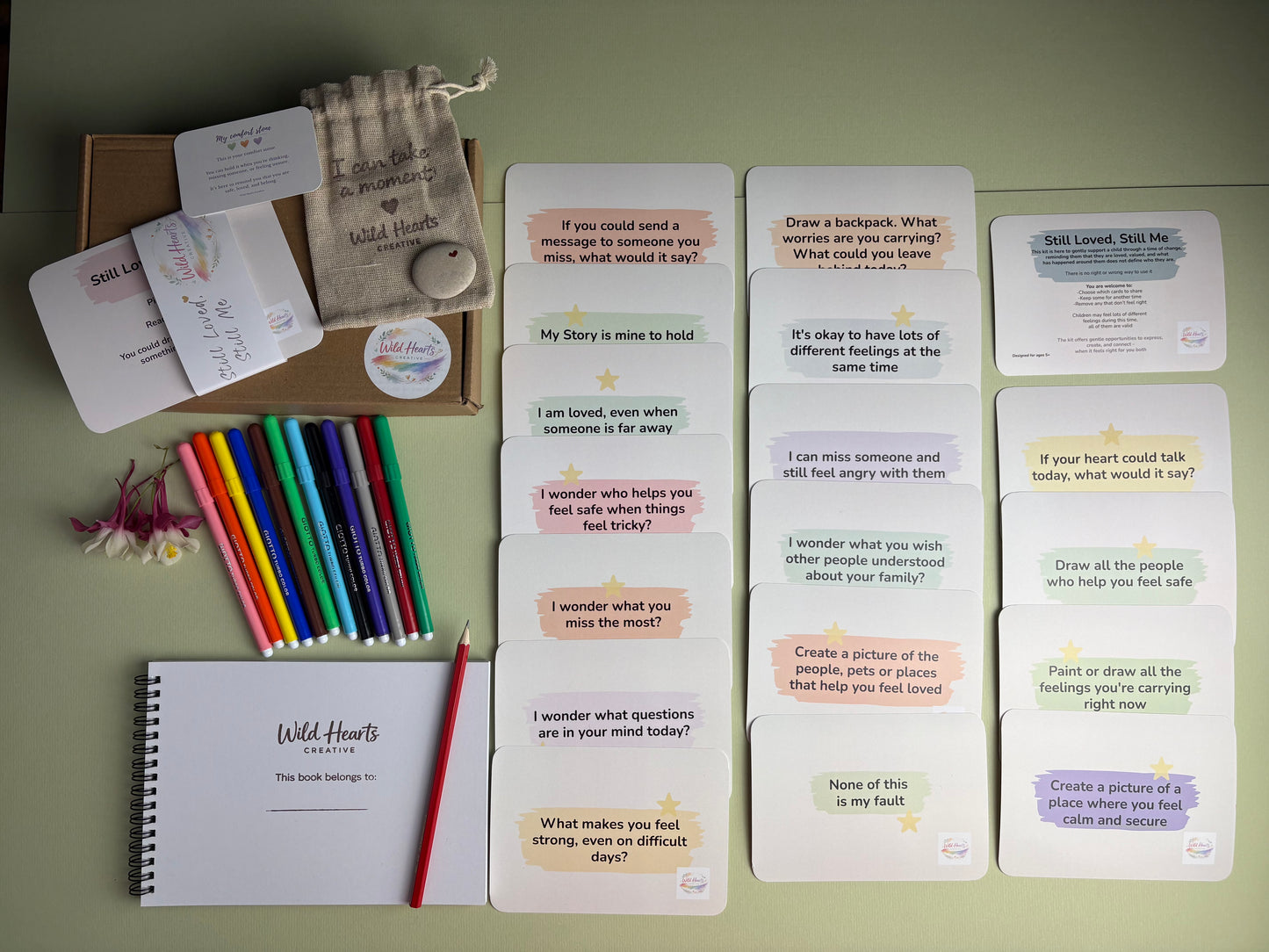 Wild Hearts Creative Still Loved, Still Me support kit displayed with reflection cards, creative activity prompts, affirmation cards, sketchbook, colouring pens, comfort stone and drawstring pouch. Designed to help children explore feelings about parental imprisonment, separation, loss, identity and connection through gentle conversation and creative activities.