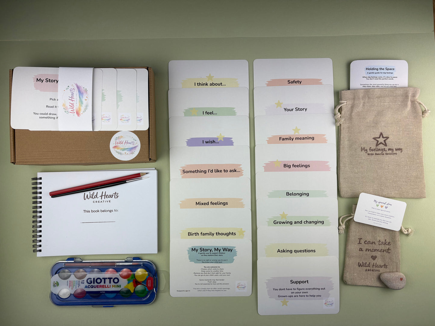Flat lay of the Adoption and Life support kit for children, including affirmation cards, creative prompt cards, sketchbook, paints, comfort stone and drawstring pouch.