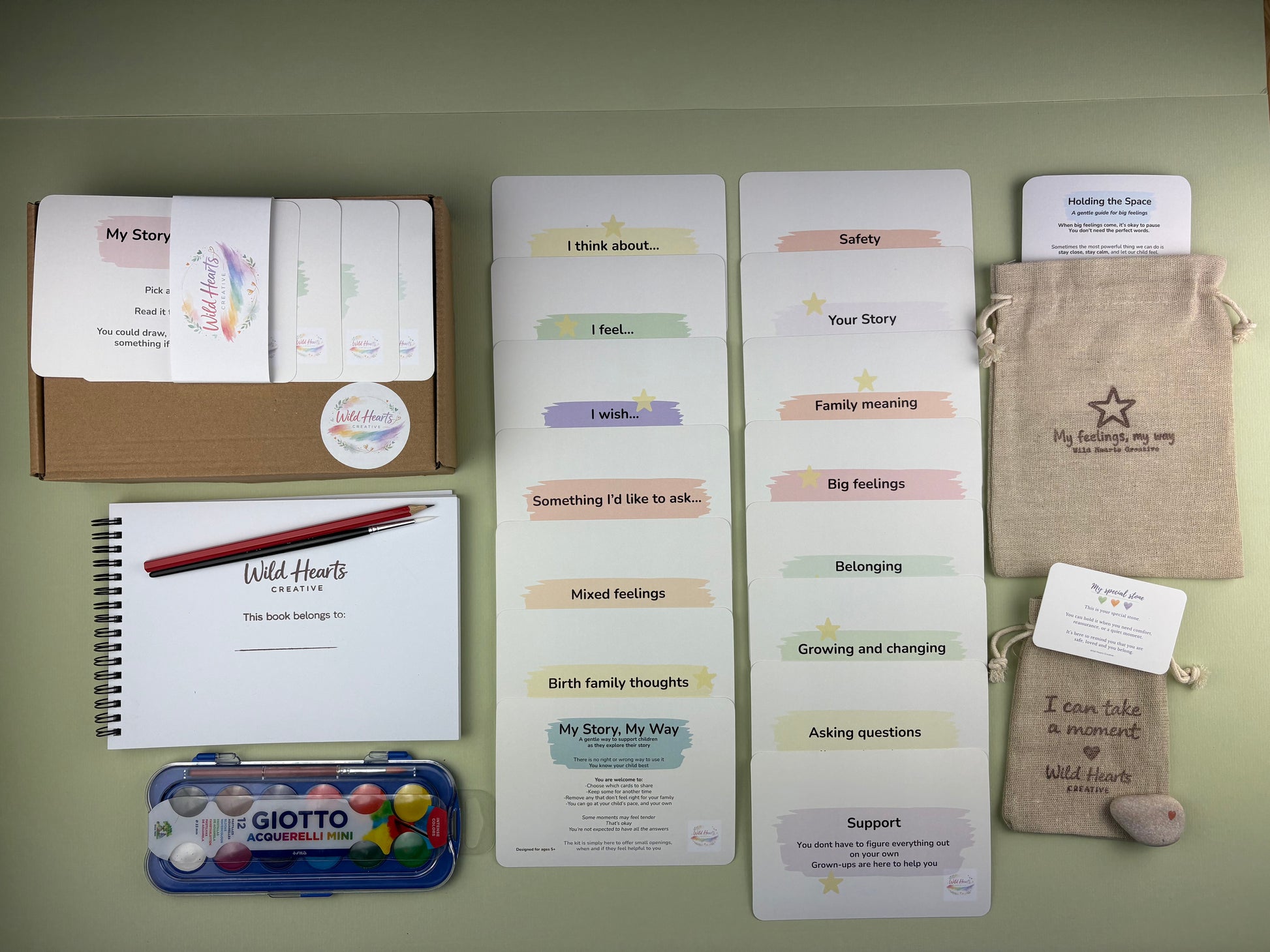 Flat lay of the Adoption and Life support kit for children, including affirmation cards, creative prompt cards, sketchbook, paints, comfort stone and drawstring pouch.