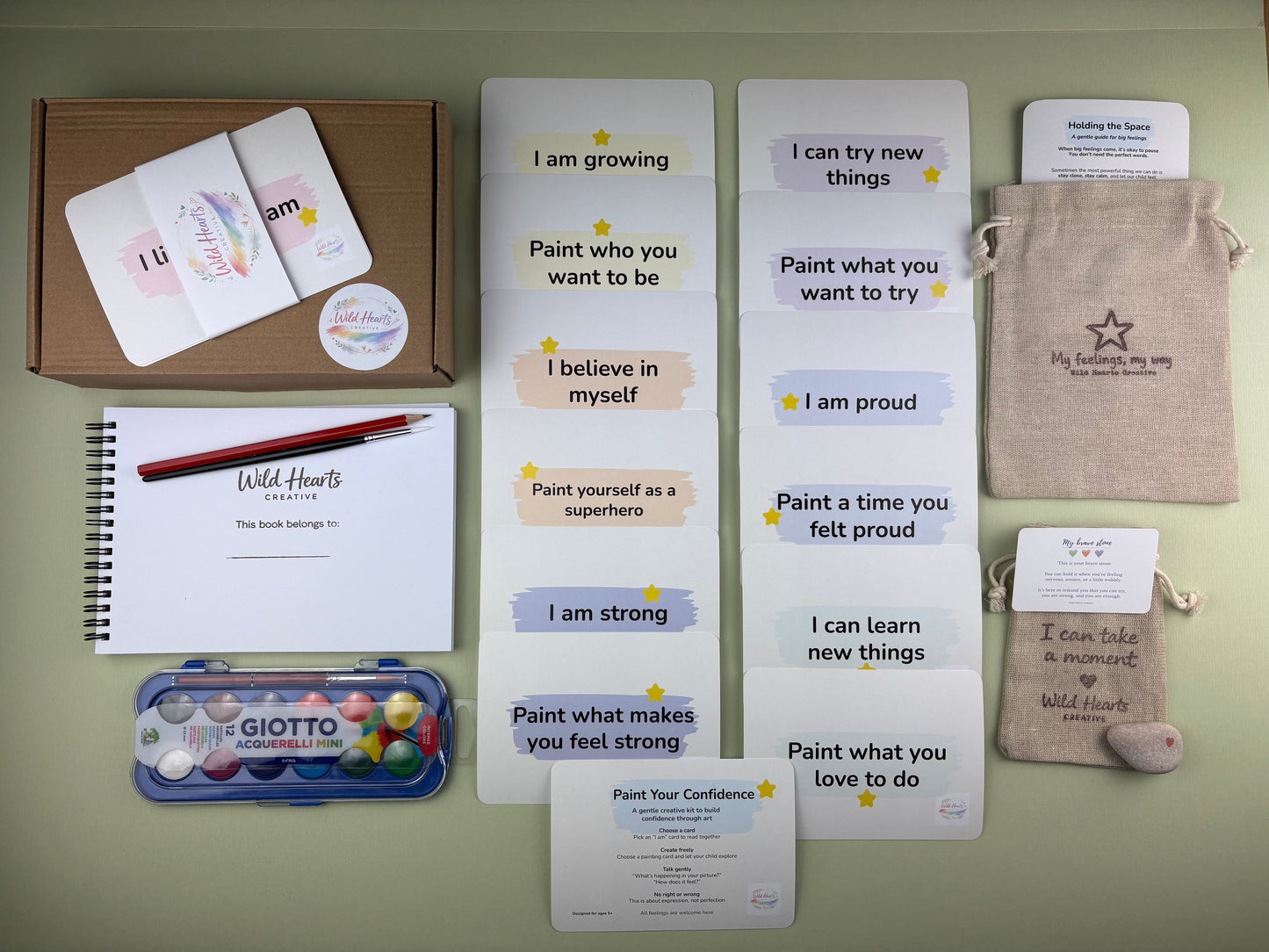 Flat lay of the Paint Your Confidence for self-belief for children, including affirmation cards, creative prompt cards, sketchbook, paints, comfort stone and drawstring pouch