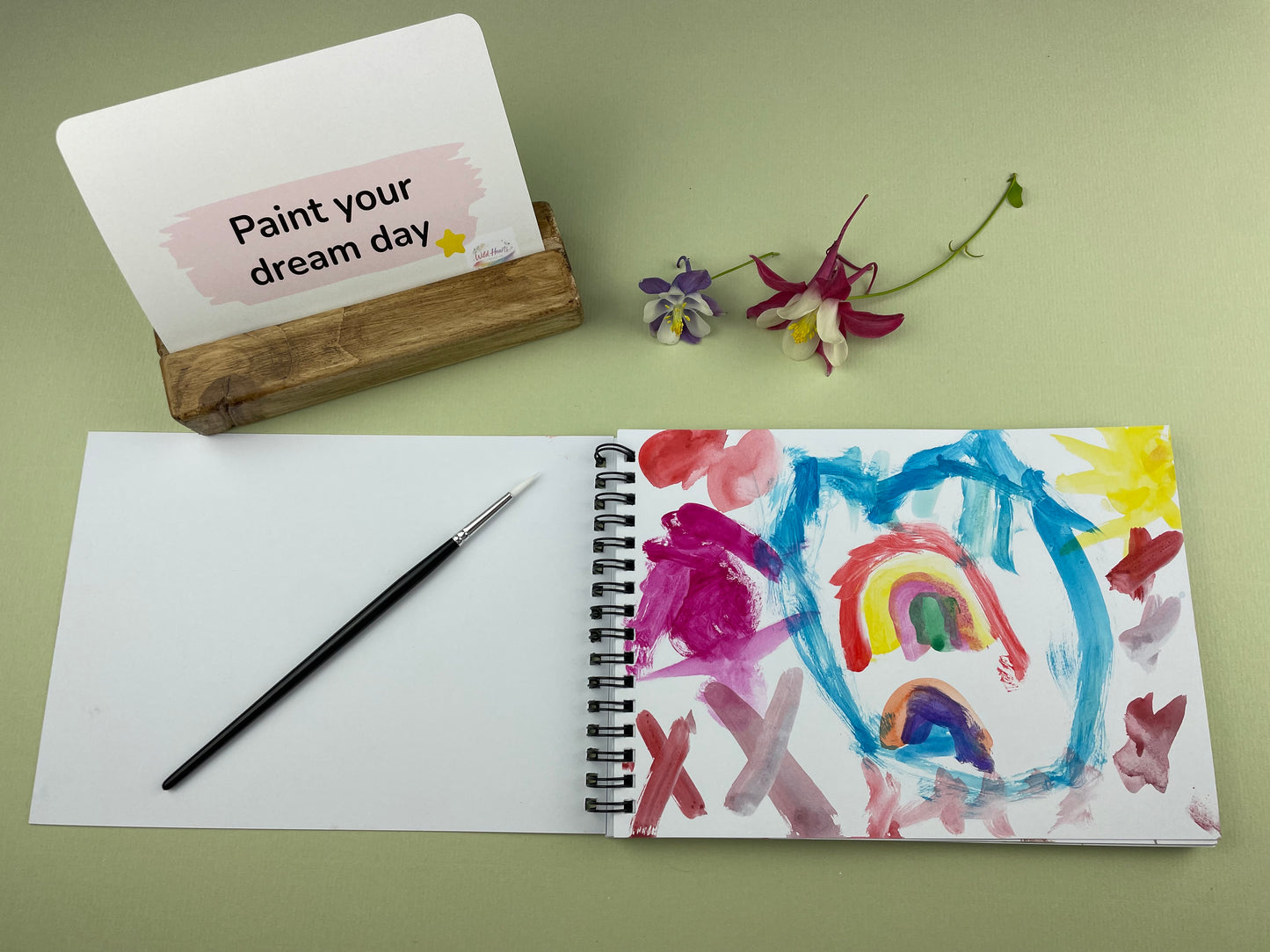 Open sketchbook showing a colourful child’s painting inspired by the Paint Your Confidence creative support kit, with prompt card, paintbrush and wooden card holder displayed on a sage green background.