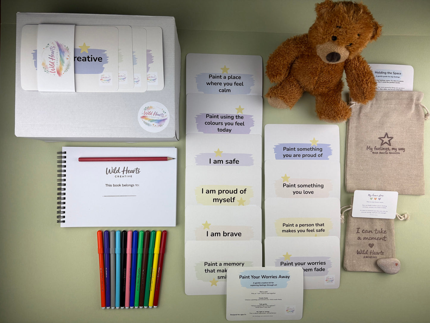 Flat lay of the Paint Your Worries Anxiety support kit for children, including affirmation cards, creative prompt cards, sketchbook, paints, comfort stone and drawstring pouch.