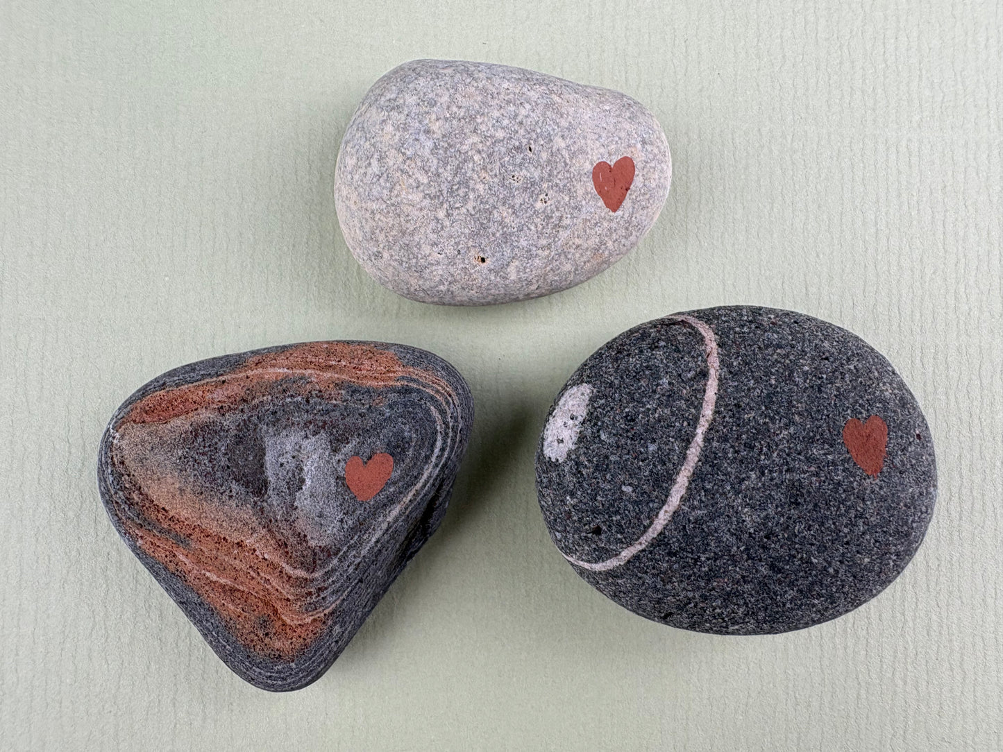 Close-up of a smooth grey comfort stone with a painted heart, grey and rust beach stone with painted heart and ring of white on grey beach stone with painted hear , designed by Wild Hearts Creative to help children feel calm, reassured and emotionally supported.