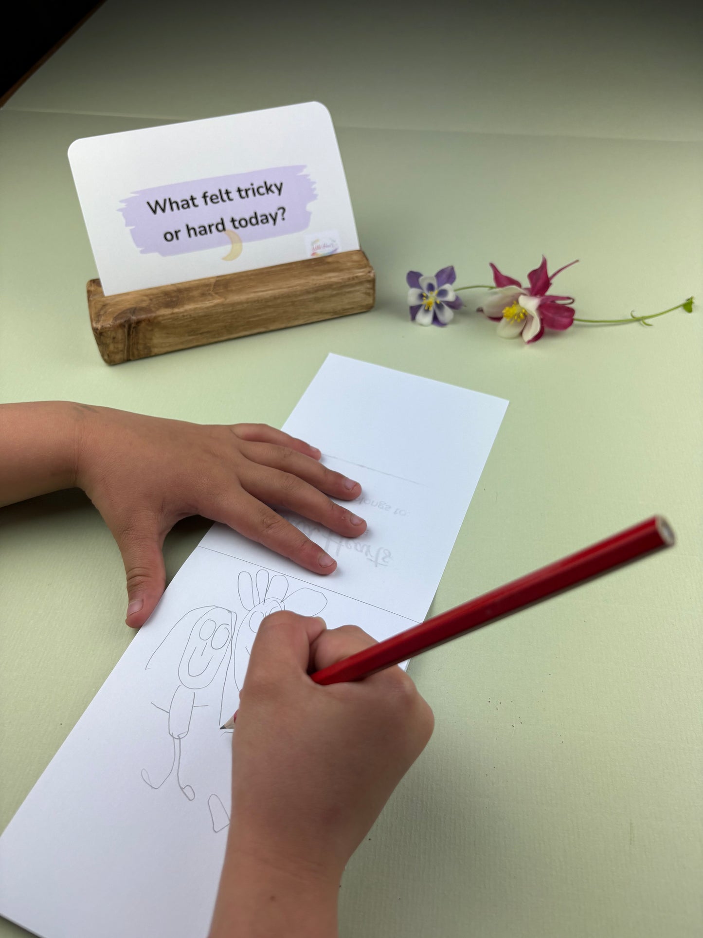 Child's hand holding a red pencil over a piece of paper with a drawing, next to a card on a light green surface.  Processing difficult emotions card, resting worries and anxious feelings at bedtime so can sleep.