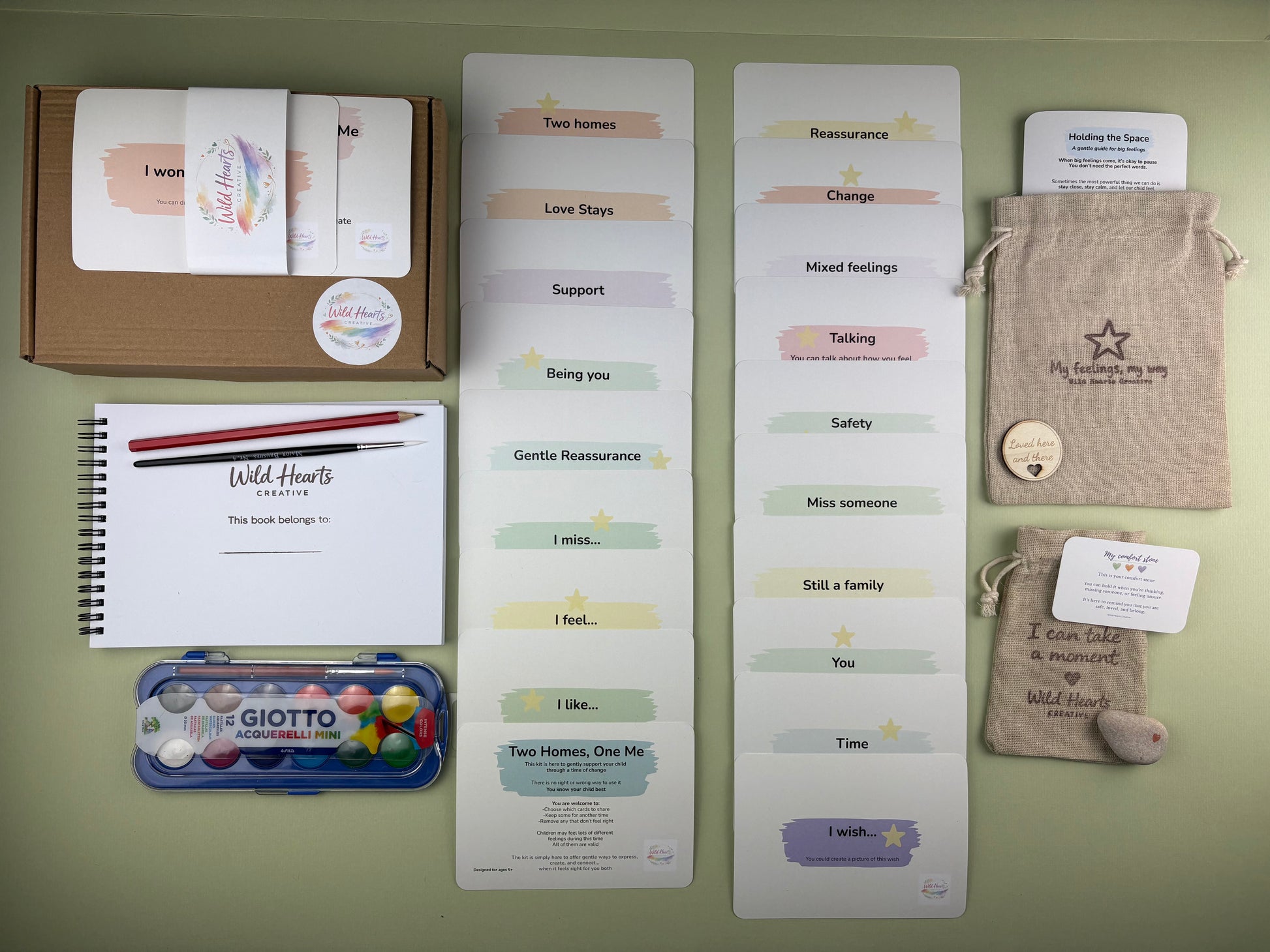 Flat lay of the Two Homes, One Me parent separation support kit for children, including affirmation cards, creative prompt cards, sketchbook, paints, comfort stone and drawstring pouch.