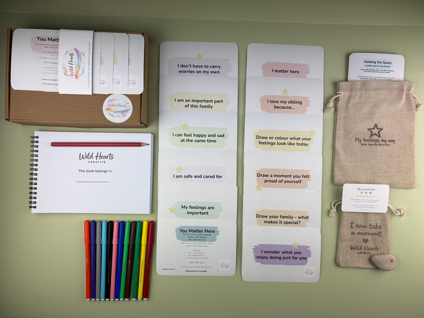 Flat lay of the You Matter Here Sibling support kit for children, including affirmation cards, creative prompt cards, sketchbook, paints, comfort stone and drawstring pouch.