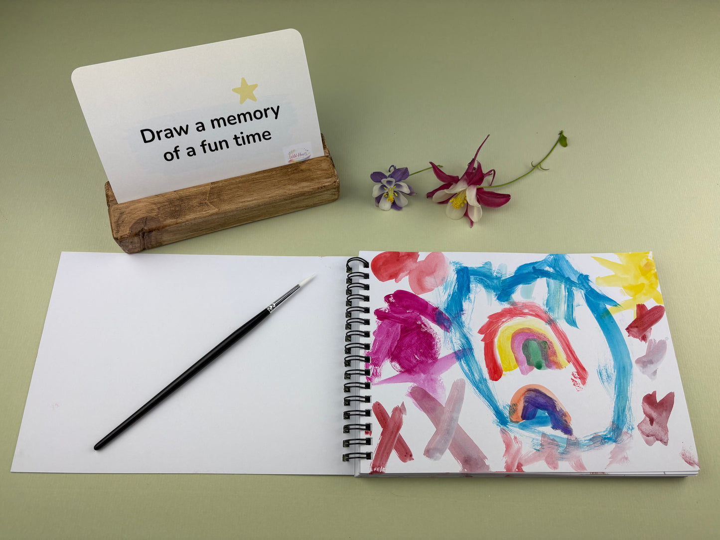 Open sketchbook showing a colourful child’s painting inspired by the Remembering You  Bereavement Grief creative support kit, with prompt card, paintbrush and wooden card holder displayed on a sage green background.