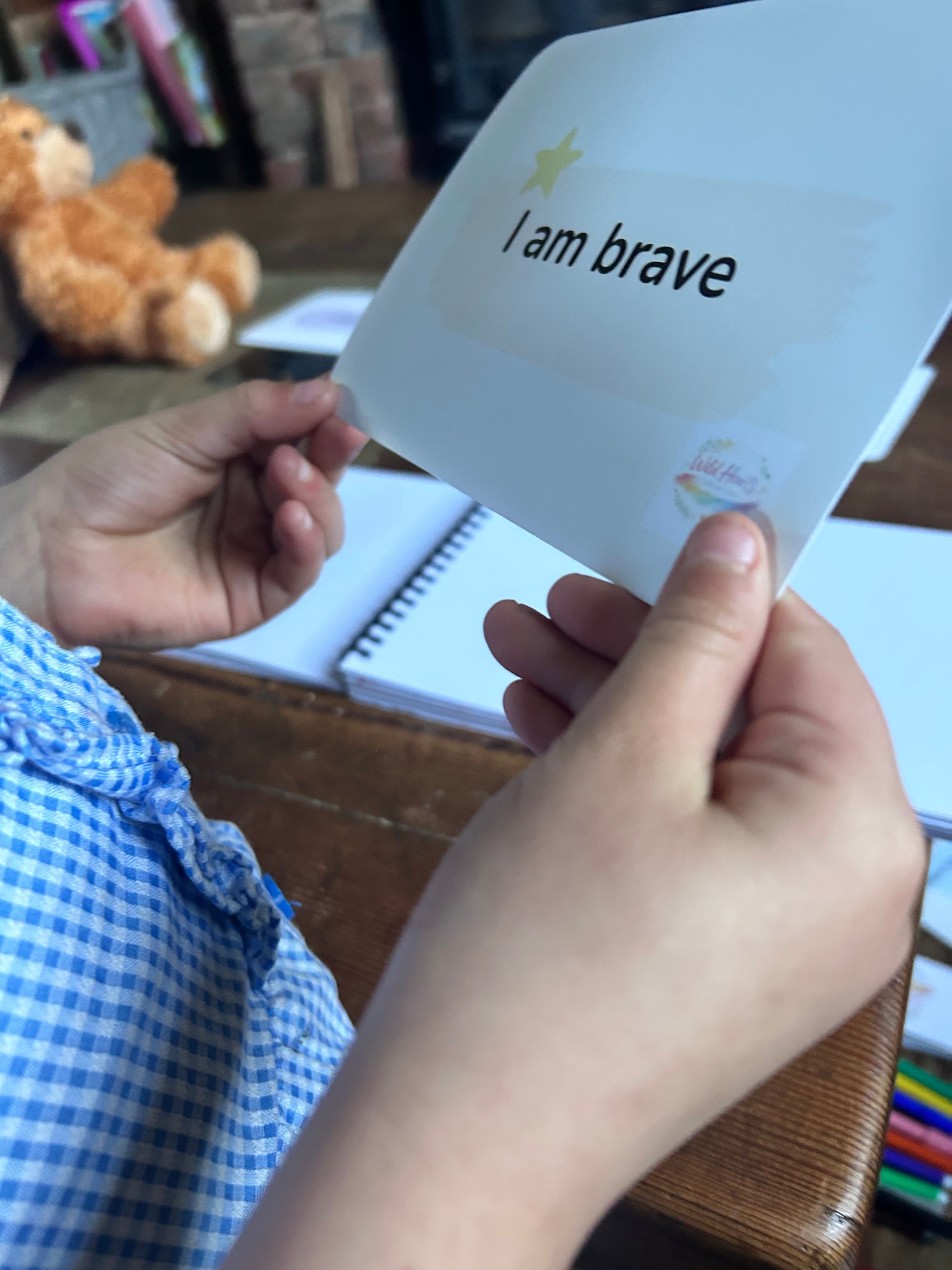 Hand holding a card with 'I am brave' affirmation card, surrounded by books and toys, branded with Wild Hearts Creative